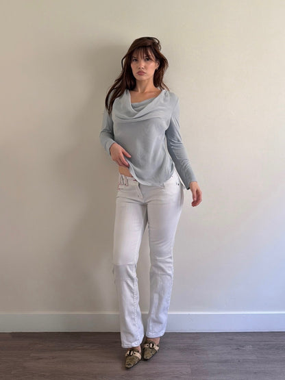 Woman wearing a light gray long-sleeve top and white jeans against a plain wall.