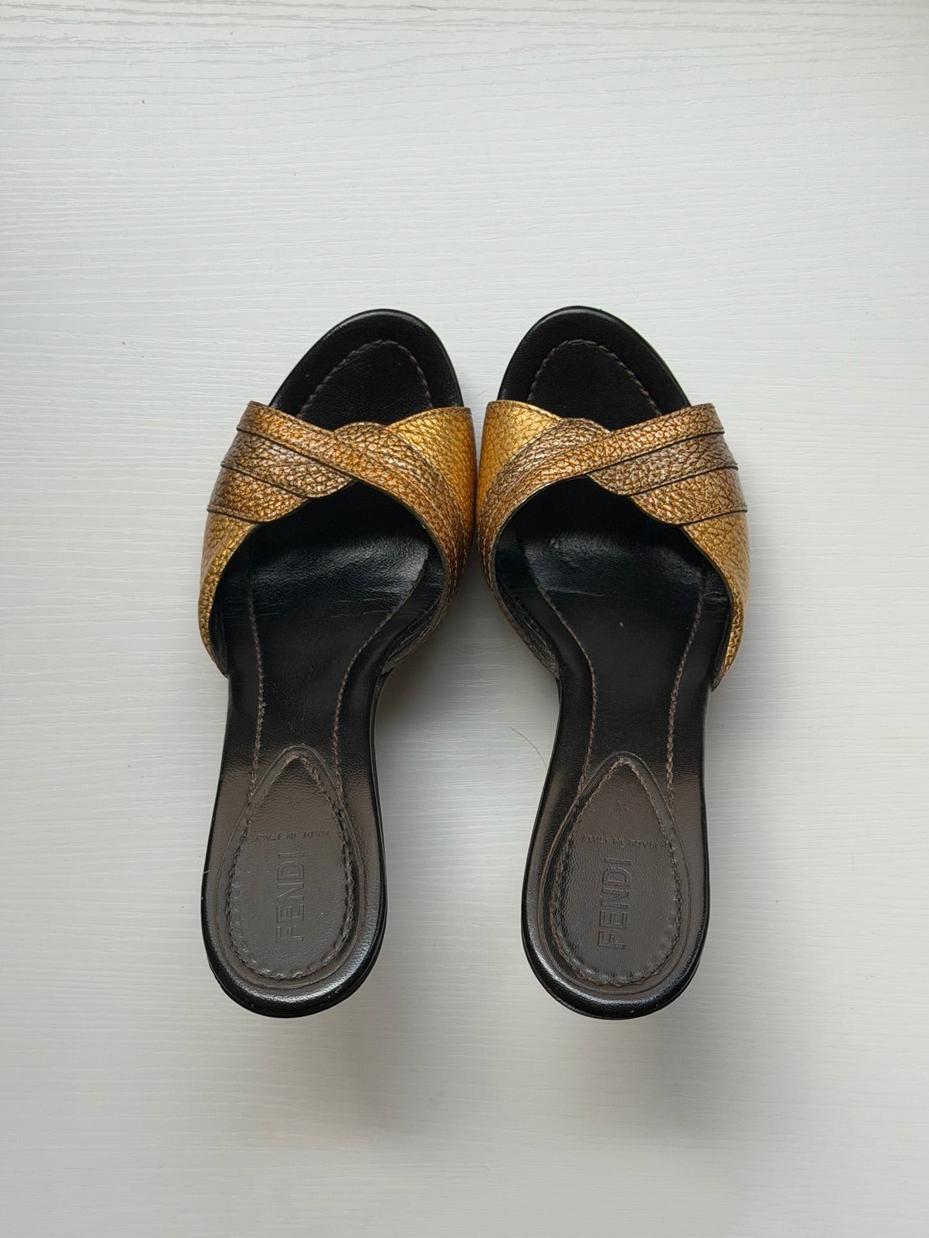 A pair of women's slide sandals with metallic gold crisscross straps and black soles, shown from a top-down view on a light gray background.