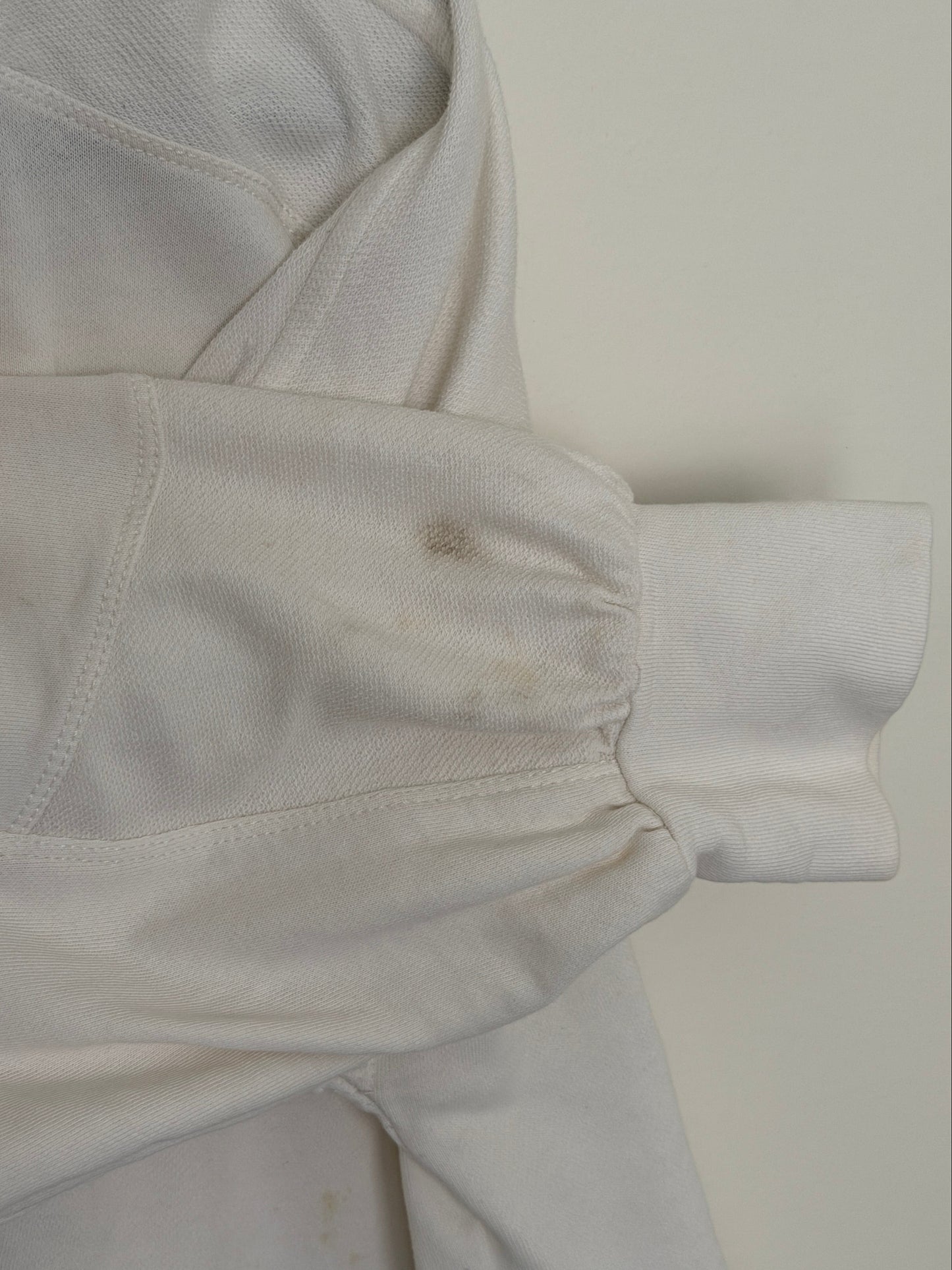 A close-up of a white long-sleeve garment laid flat, showing the sleeve cuff, stitched seam, and a visible hand holding the fabric. The garment appears slightly wrinkled and has some discoloration or stains.