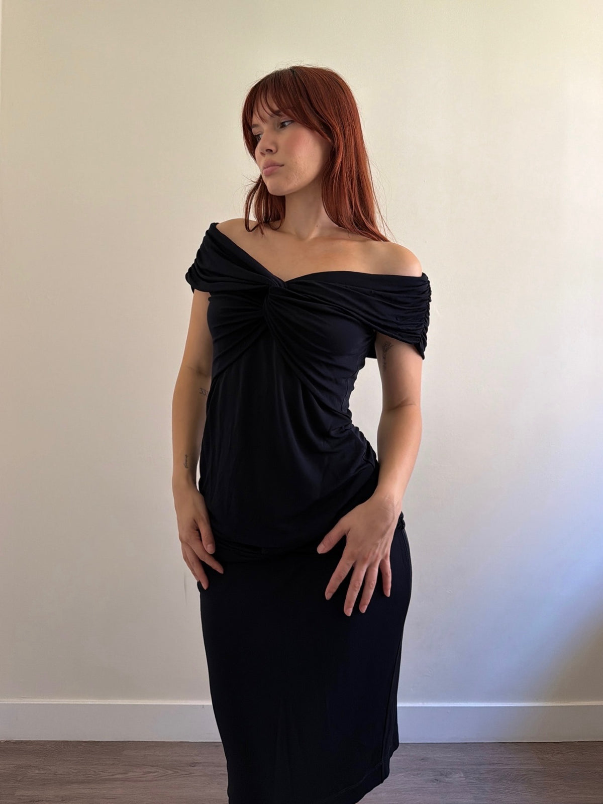A woman with red hair wearing a black off-the-shoulder midi dress, standing against a plain off-white wall.