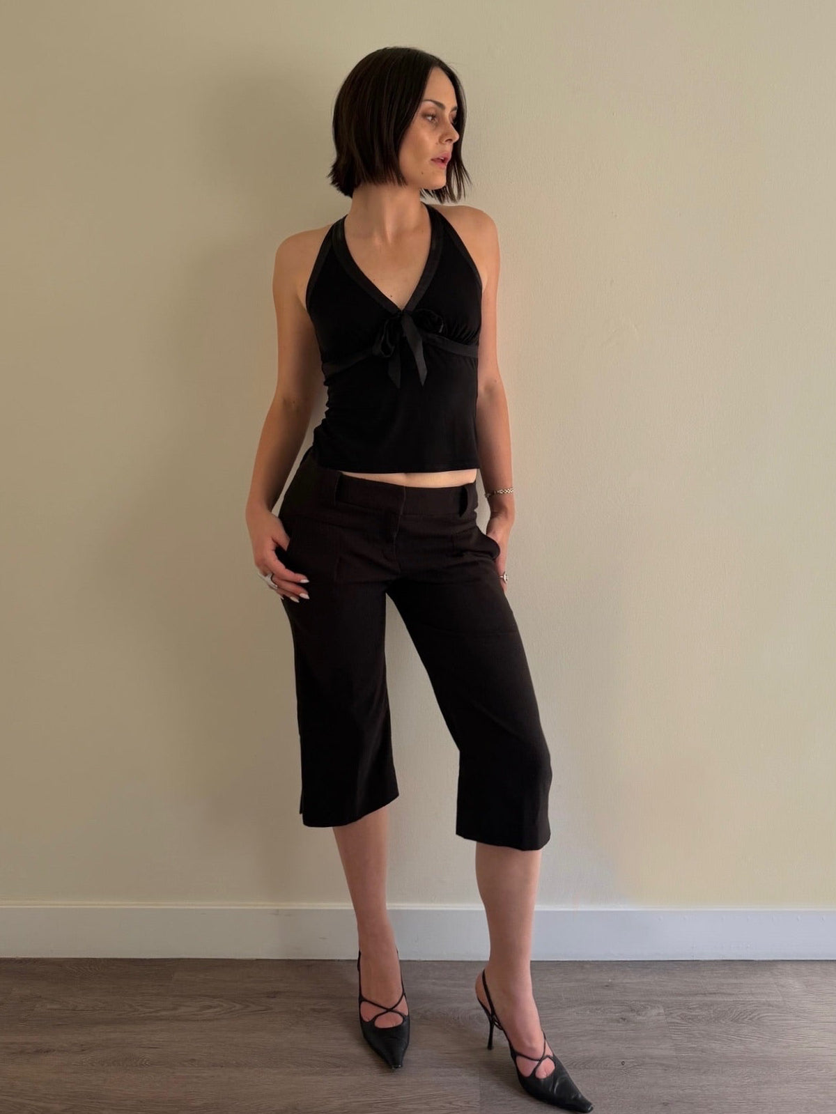 Woman wearing a black halter top and black capri pants against a beige wall.