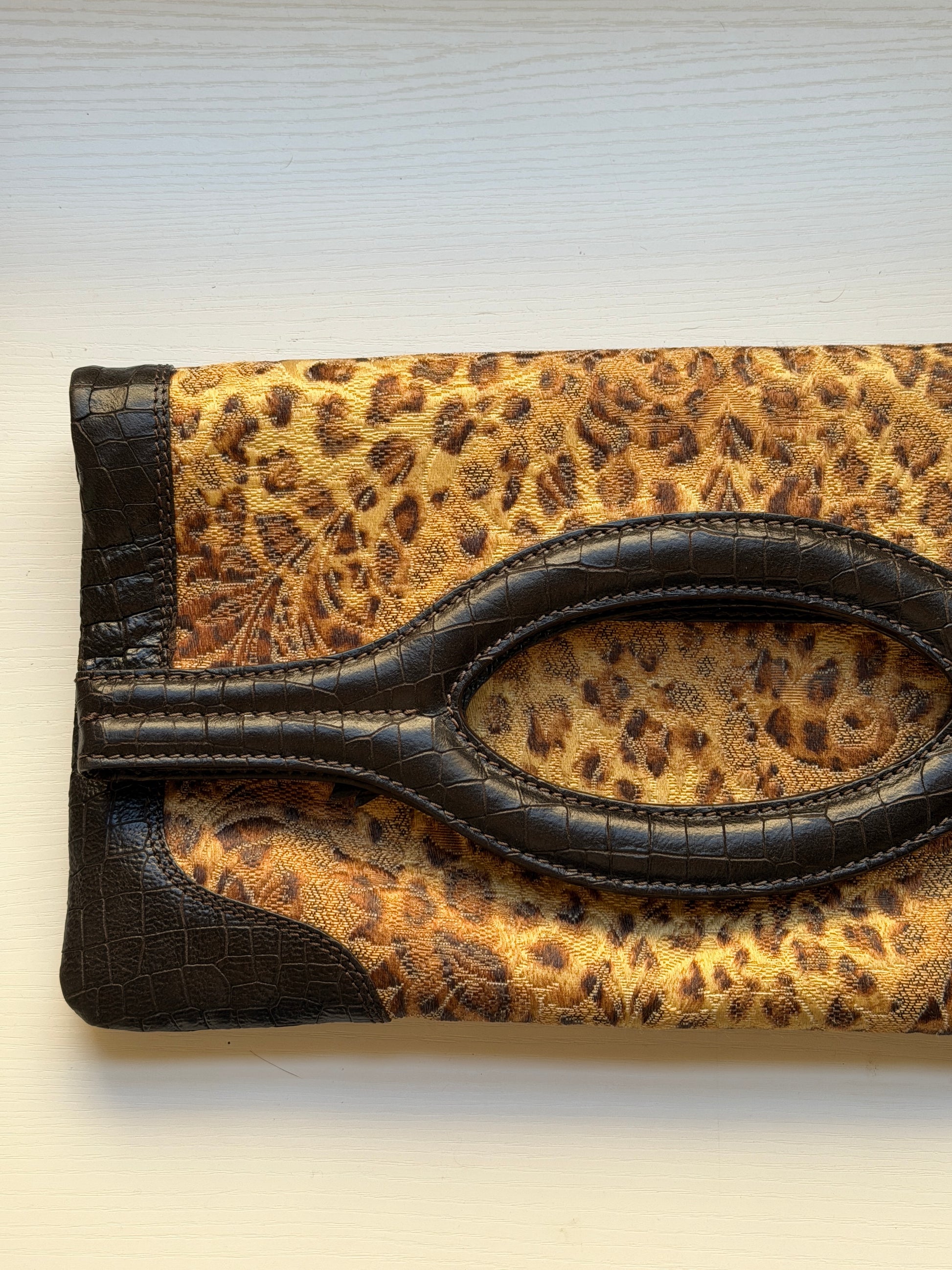 A leopard print clutch purse with dark brown crocodile-embossed trim and a large oval handle cutout, shown on a light wood background.