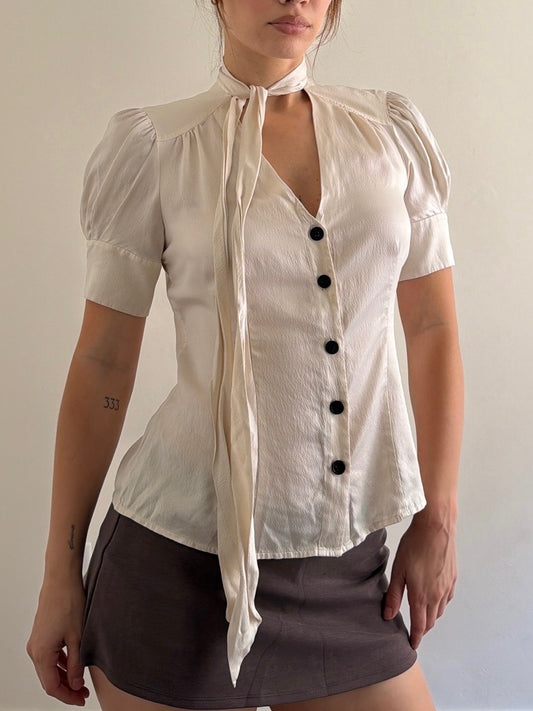 Woman wearing a white blouse with black buttons and a brown skirt against a plain background