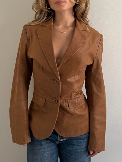 A woman wearing a fitted tan-brown leather blazer with notched lapels and two front buttons, paired with blue jeans, shown against a plain light wall.