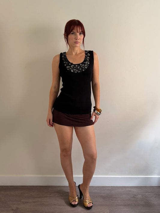 A woman stands against a plain beige wall wearing a black sleeveless top with a dark gray floral pattern around the neckline, a dark brown mini skirt, and gold high-heeled sandals.