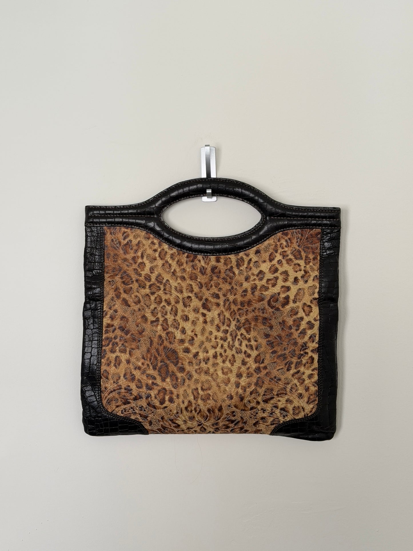 A leopard-print handbag with black faux-croc trim and a top handle, hanging from a small metal hook against a plain light gray wall.