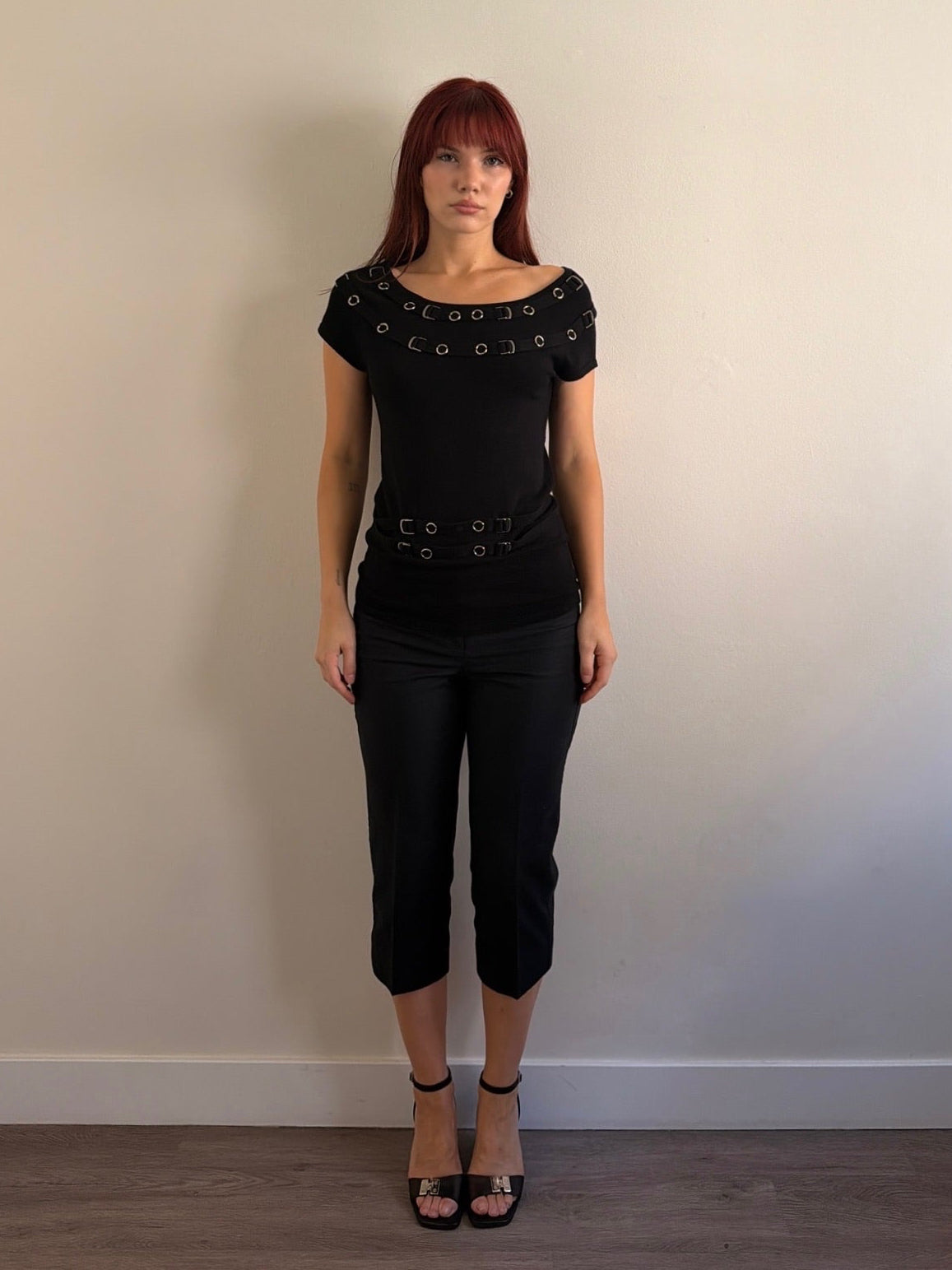 Woman wearing a black top with decorative elements and black pants against a plain wall.