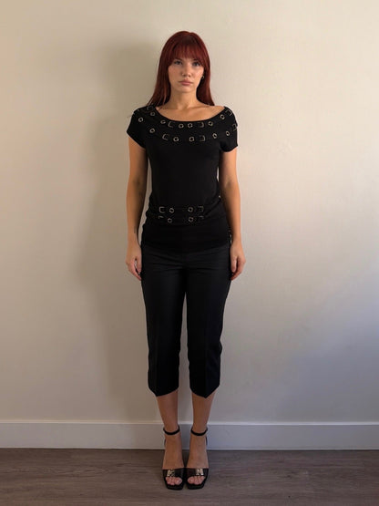Woman wearing a black top with decorative elements and black pants against a plain wall.
