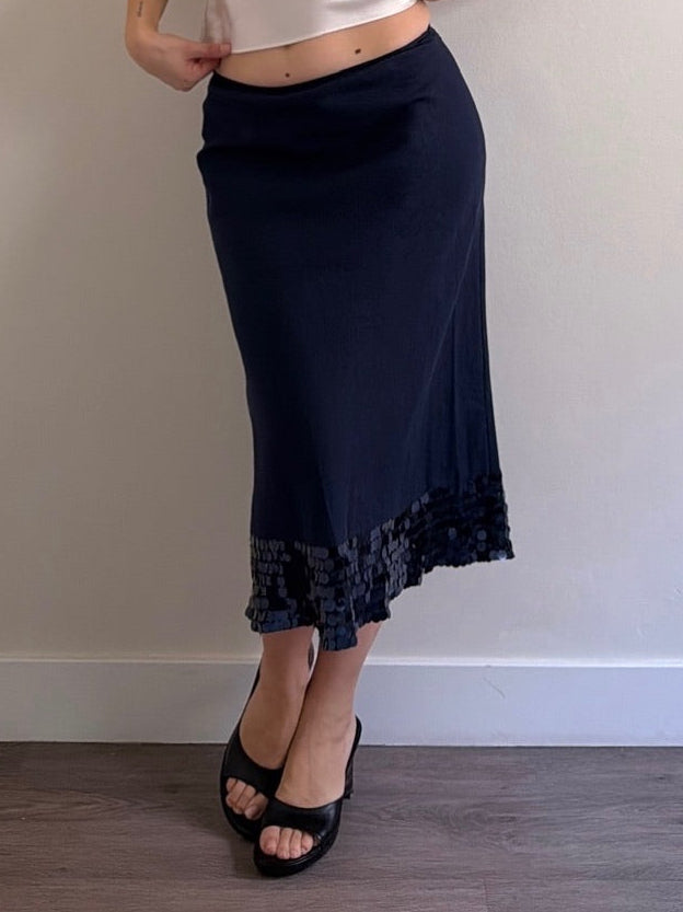 Woman wearing a white top and navy skirt against a plain wall.