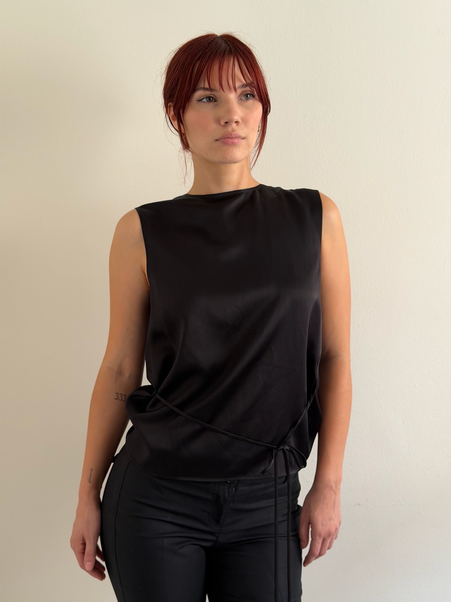 A woman with red hair wearing a sleeveless black satin top with a wrap-style tie detail at the waist, paired with black pants, standing against a plain off-white background.