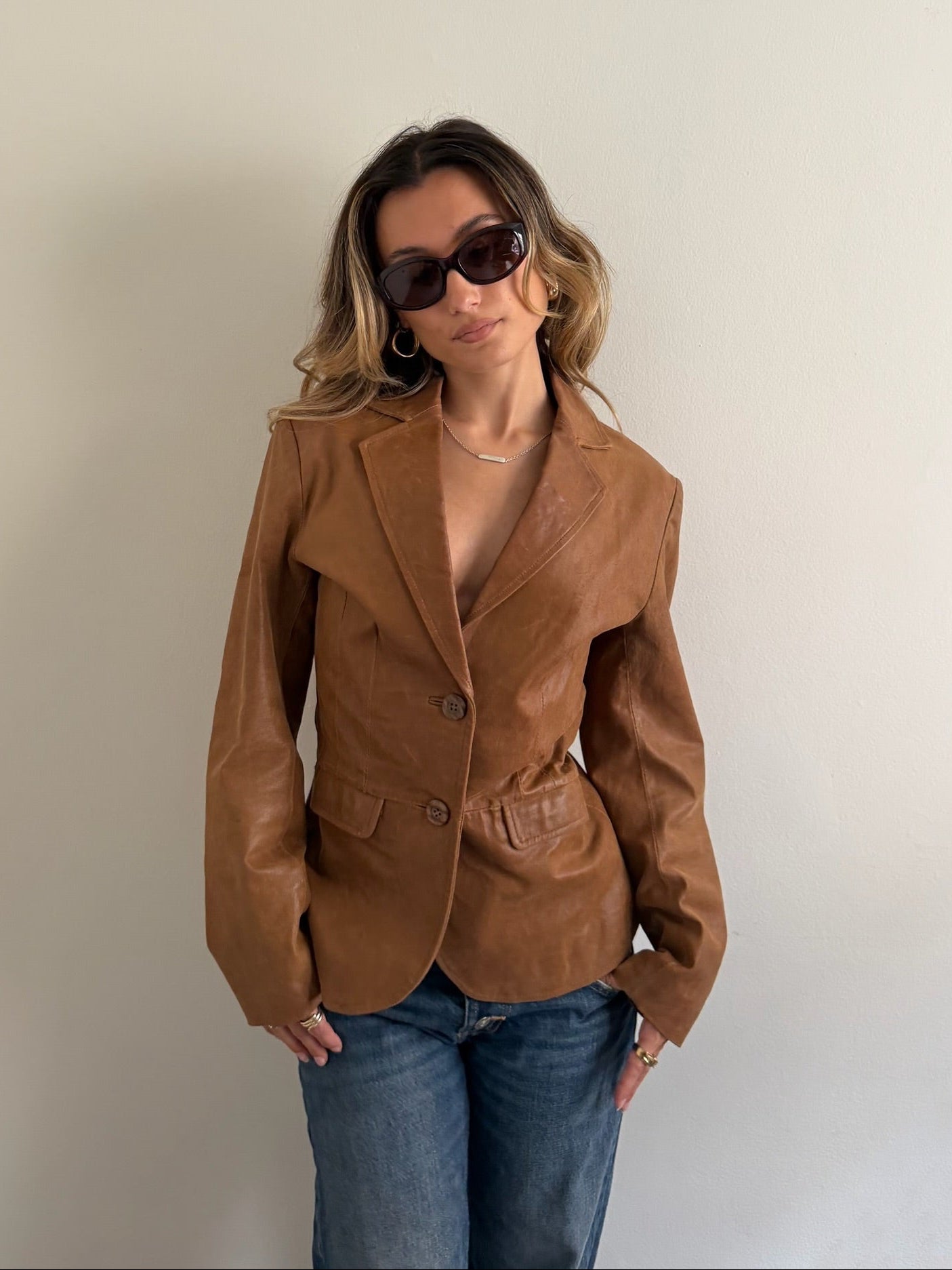 A woman wearing a camel brown faux leather blazer and blue jeans, standing against a plain off-white wall.