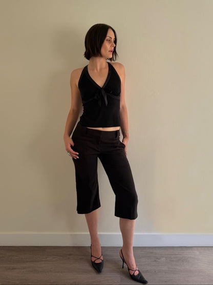 Woman wearing a black halter top and black capri pants against a beige wall.