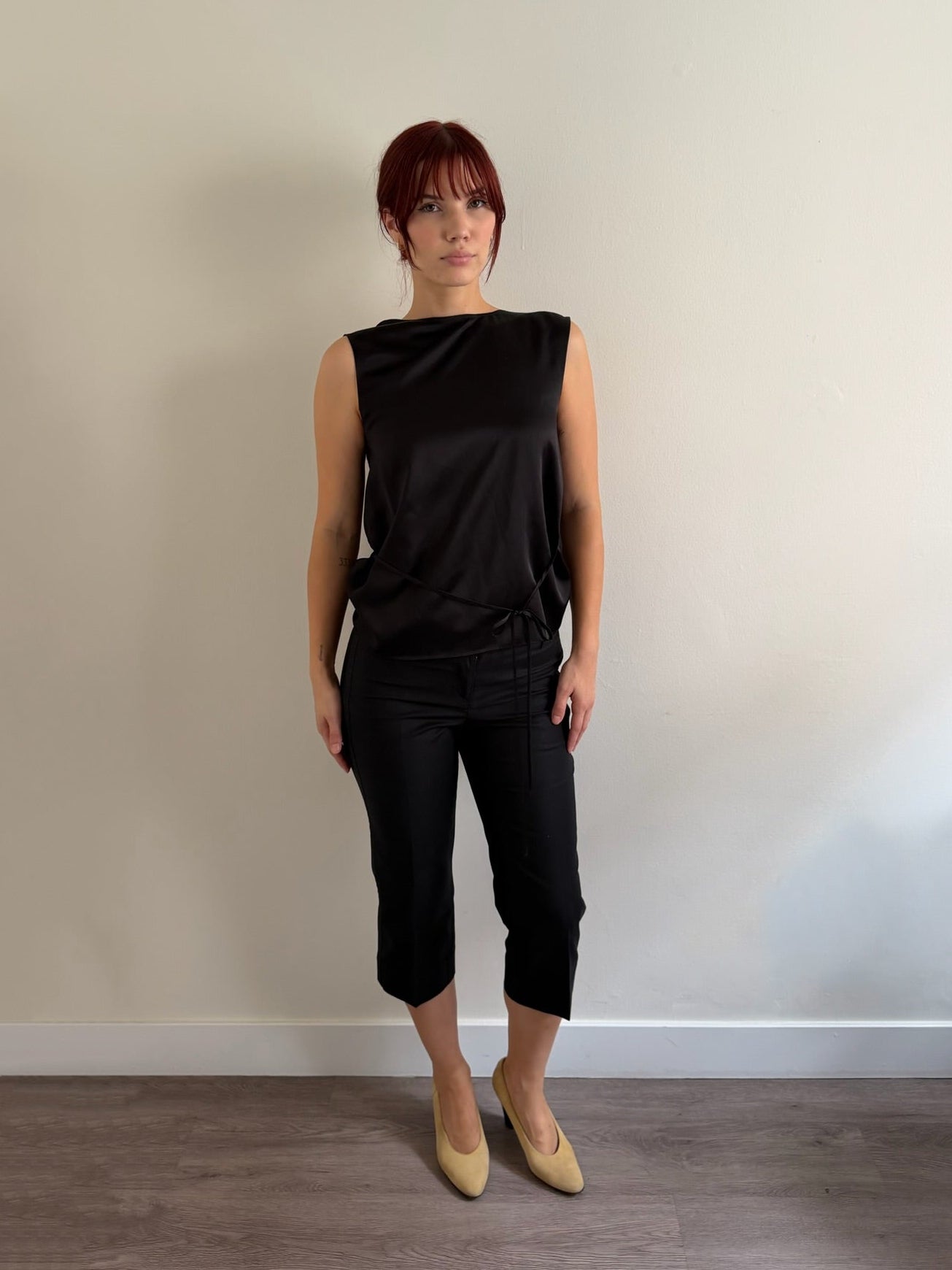 A woman with short red hair stands against a plain beige wall wearing a black sleeveless top and black cropped pants, paired with beige pointed-toe flats.