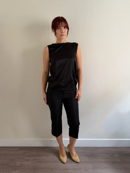 A woman with short red hair stands against a plain beige wall wearing a black sleeveless top and black cropped pants, paired with beige pointed-toe flats.
