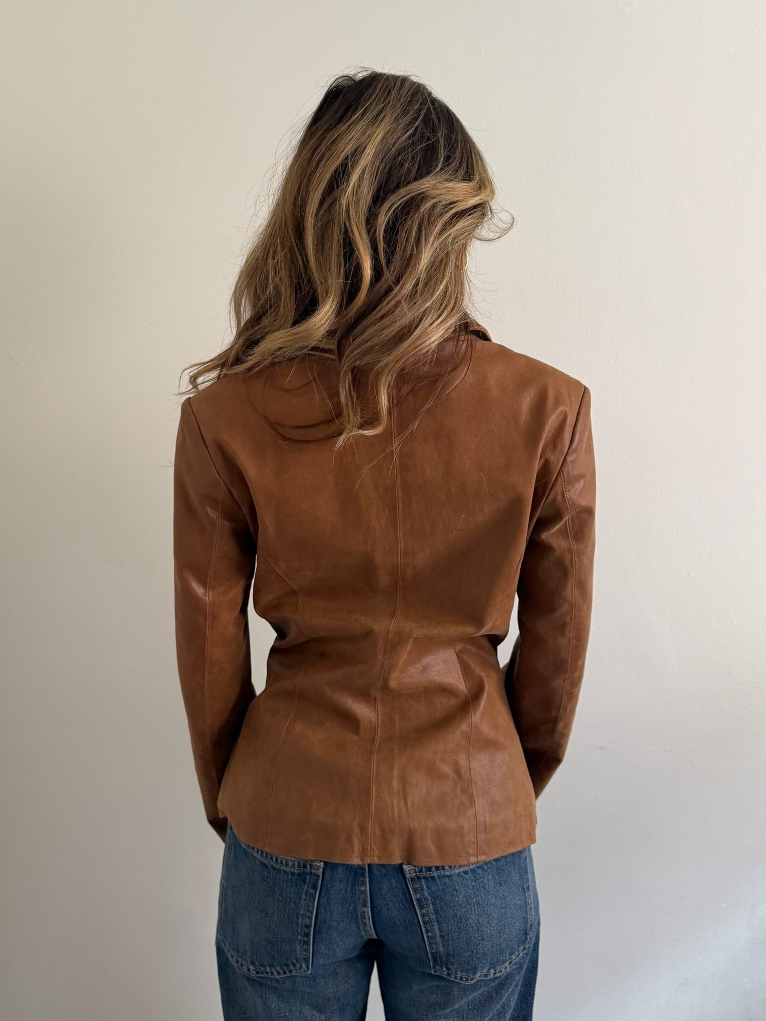 A woman viewed from behind wearing a brown faux-leather blazer and blue jeans, standing against a plain off-white wall.