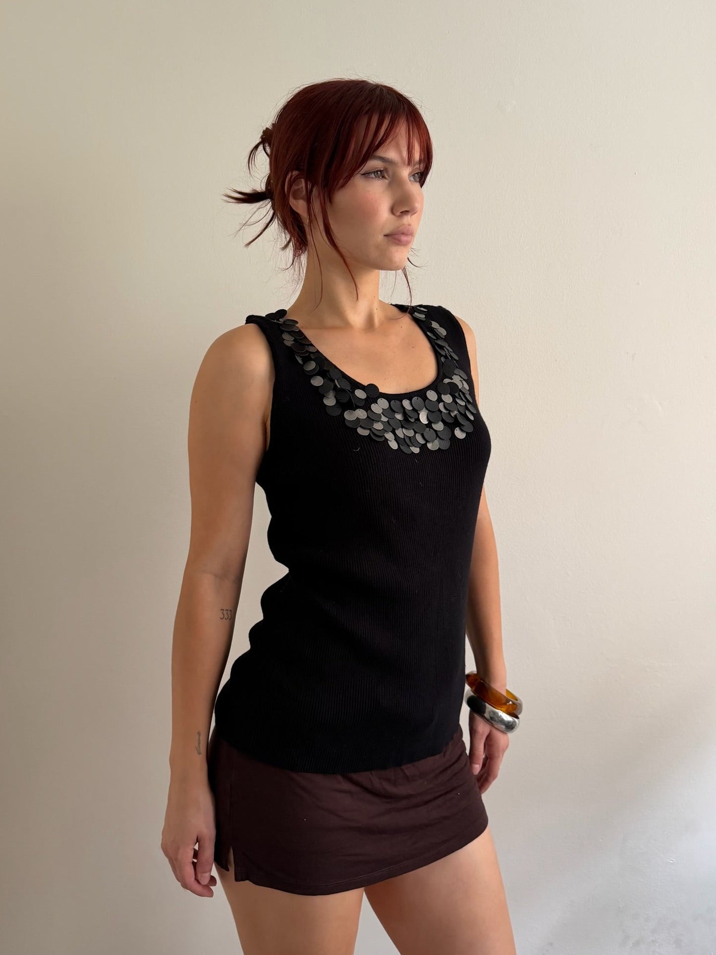 A woman with red hair wearing a black sleeveless top with a decorative patterned neckline and a dark brown mini skirt, standing against a plain off-white wall.