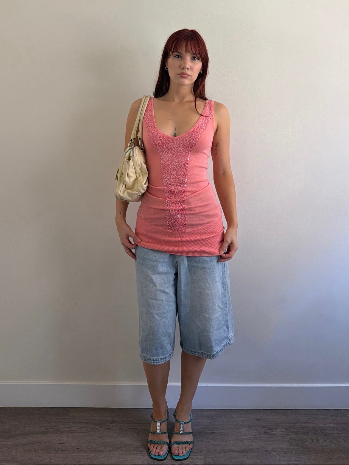 Woman wearing a pink sleeveless top and denim shorts against a plain wall.
