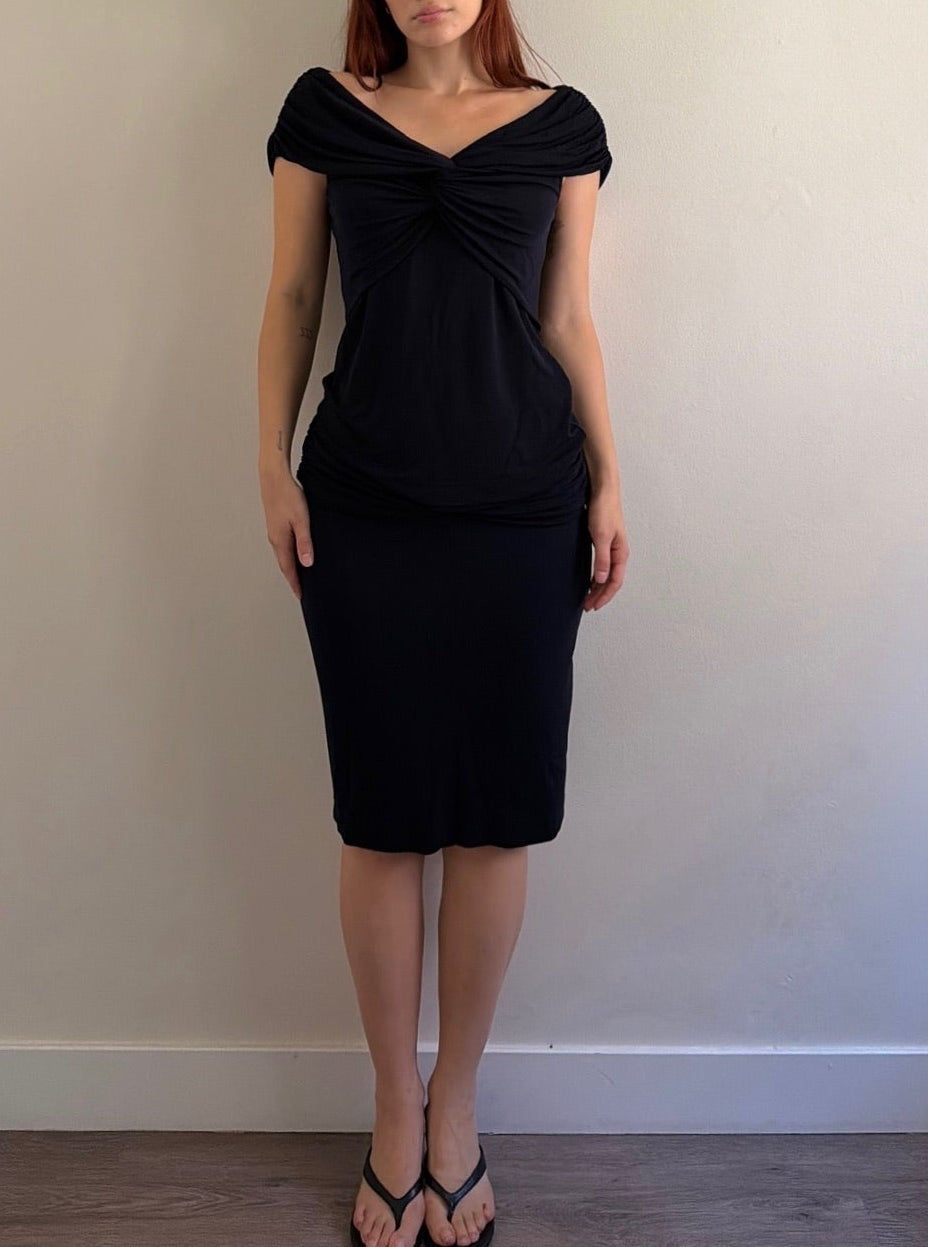 A woman with red hair wearing a black off-the-shoulder knit dress with a twisted detail at the neckline, standing against a plain beige wall.