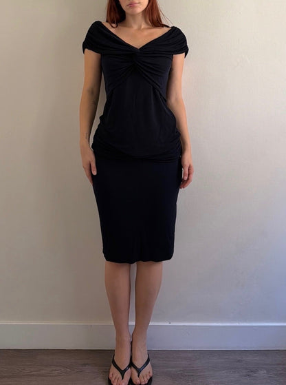 A woman with red hair wearing a black off-the-shoulder knit dress with a twisted detail at the neckline, standing against a plain beige wall.