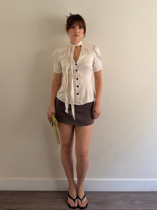 Woman wearing a white blouse with black buttons and a brown skirt against a plain wall.