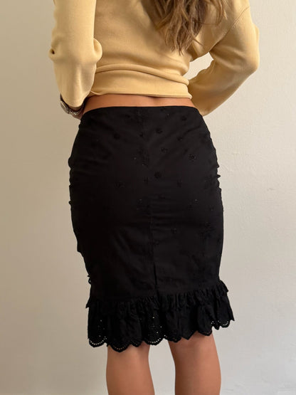 A person shown from the back wearing a black fitted pencil skirt with a ruffled hem and a beige cropped long-sleeve top, standing against a plain light wall.