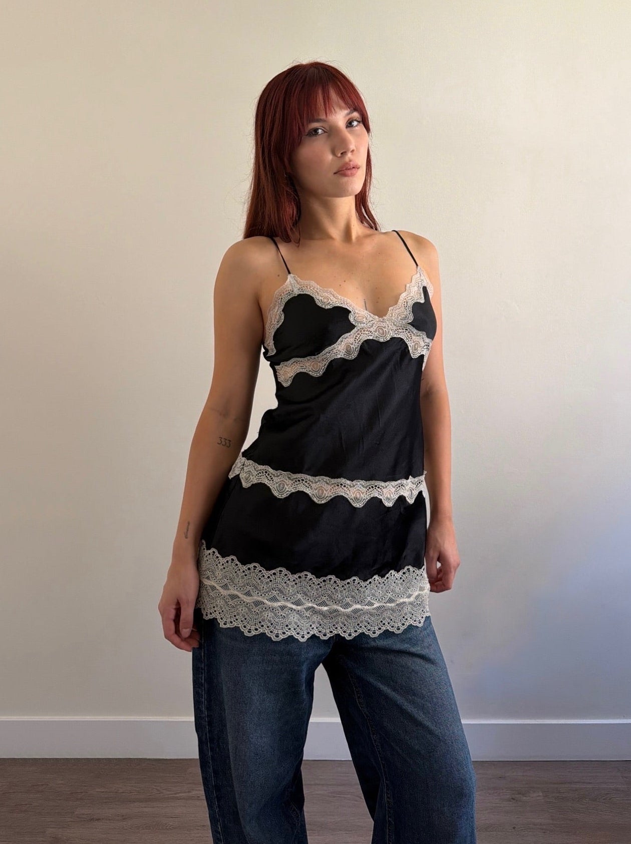 Woman wearing a black lace top and blue jeans against a plain wall.