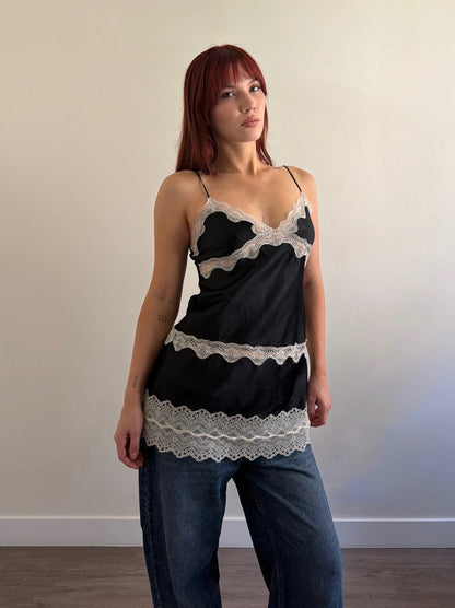 Woman wearing a black lace top and blue jeans against a plain wall.