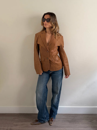 A woman wearing a tan leather blazer, blue jeans, and sunglasses stands against a plain beige wall on a wooden floor.