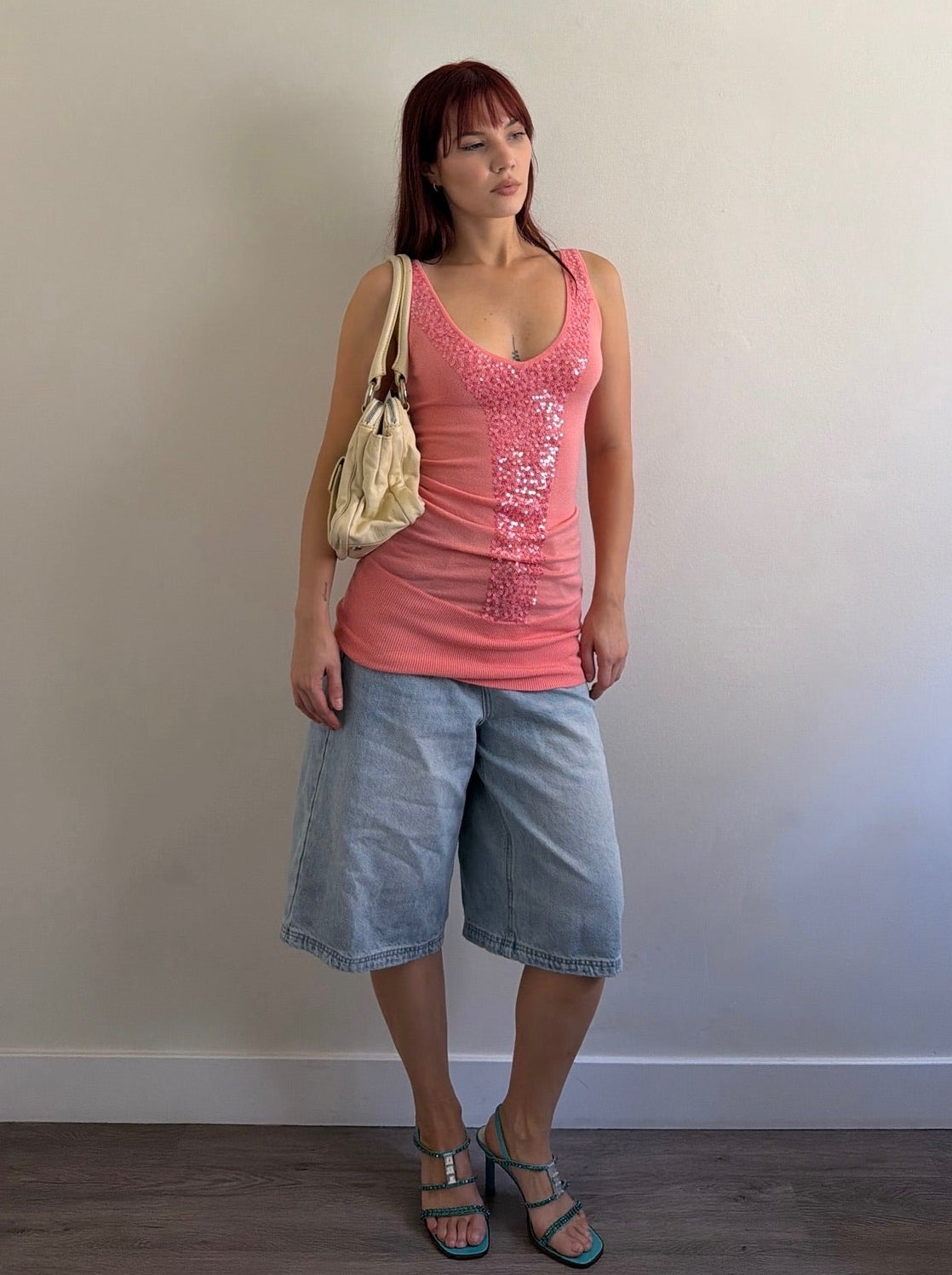Woman wearing a pink tank top and denim shorts standing against a plain wall.