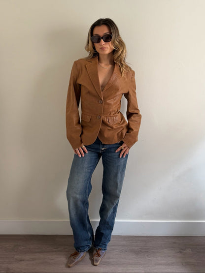 A woman wearing a tan leather blazer, blue jeans, and brown shoes stands against a plain beige wall.