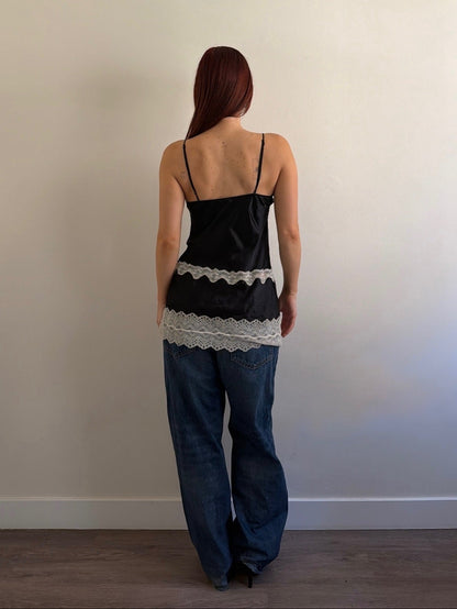 Woman wearing a black top with lace details and blue jeans against a plain wall.