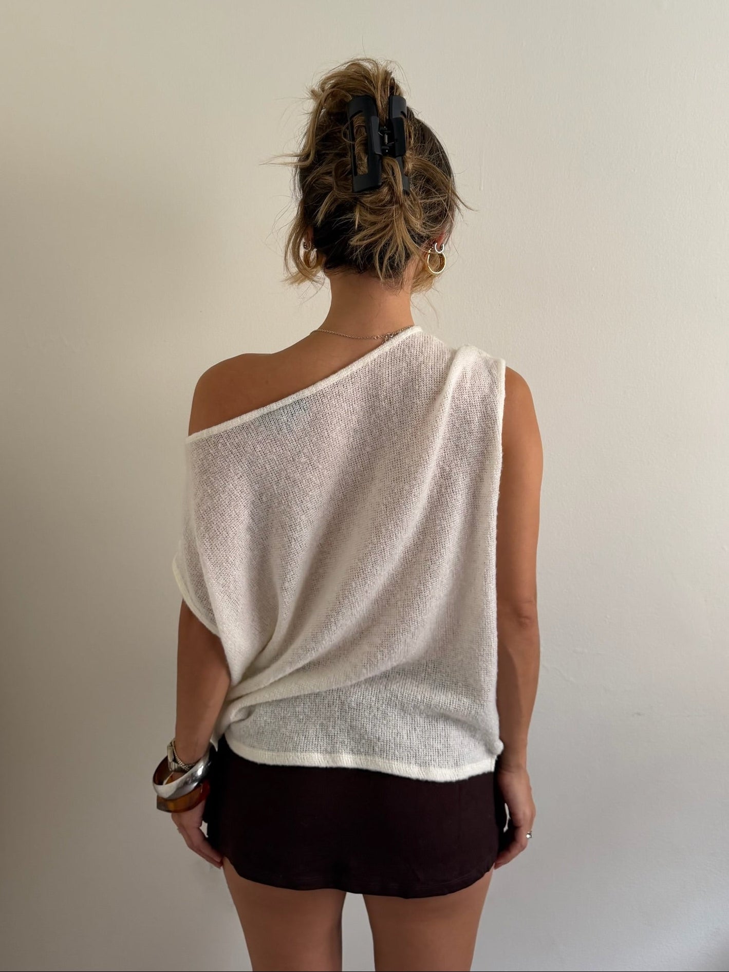A woman viewed from behind wearing a textured off-white one-shoulder knit top and dark brown shorts against a plain light gray wall.