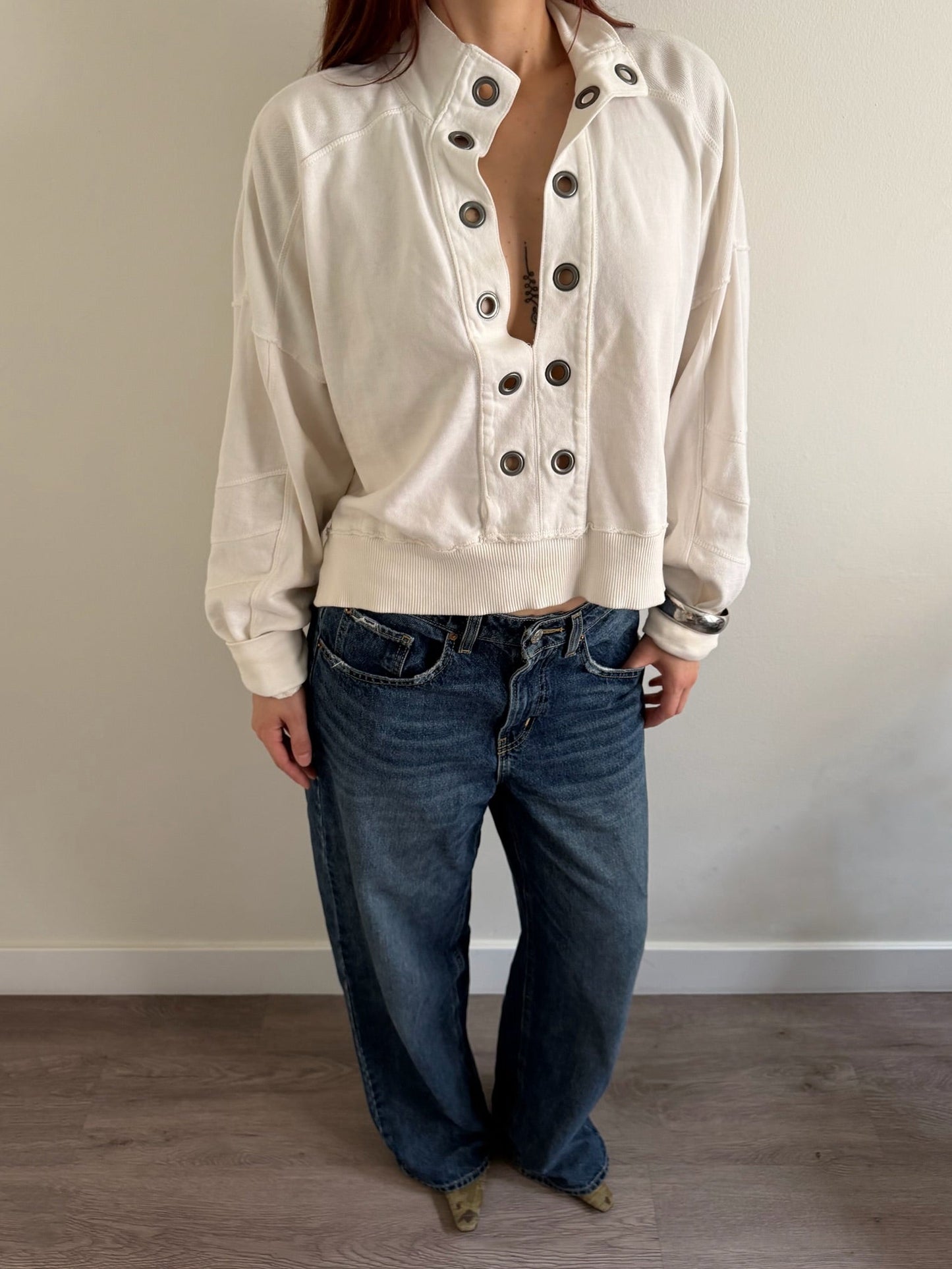A person wearing a cream-colored, cropped button-up jacket with dark buttons and blue wide-leg jeans, standing against a plain beige wall and wood floor.