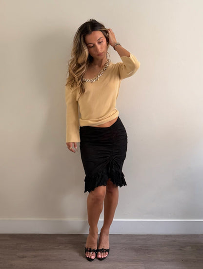 A woman modeling a pale yellow long-sleeve knit top with a pearl-like collar, paired with a black ruched midi skirt and black strappy heels, standing against a plain beige wall.