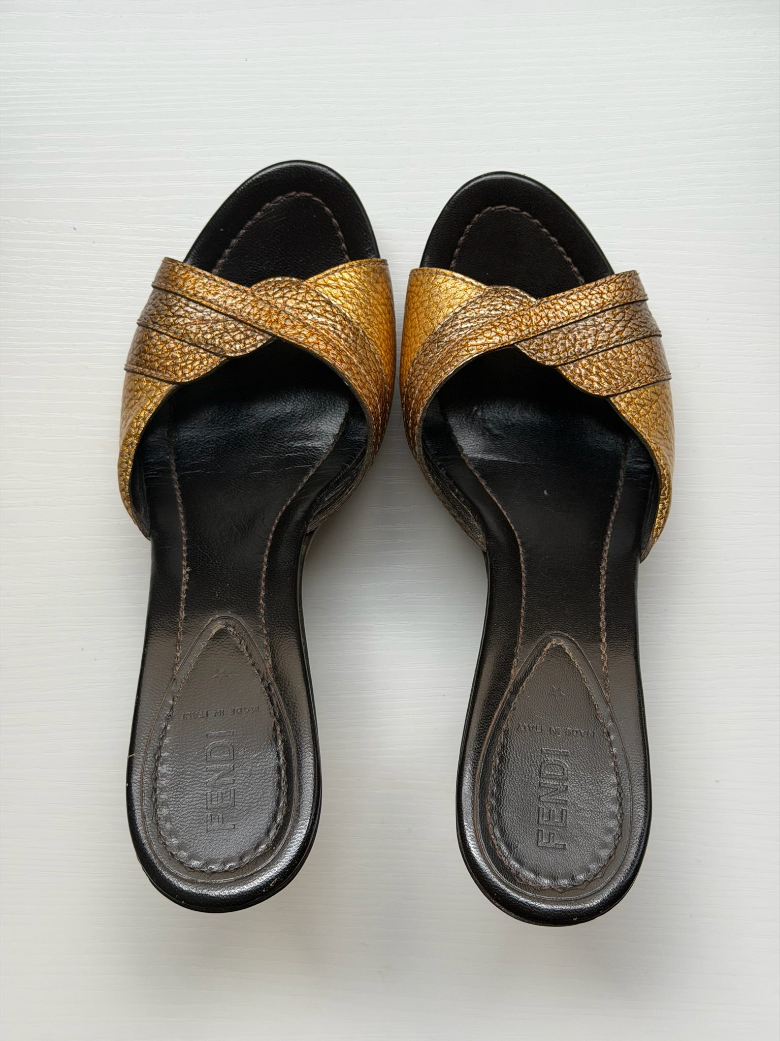 A pair of Fendi slide sandals with metallic gold woven straps and black wedge heels, shown from a top-down view on a light gray background.