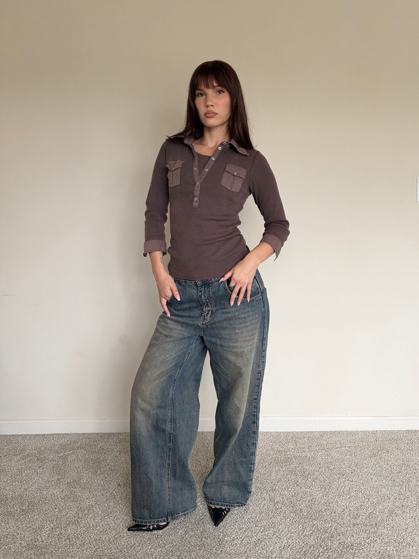 Woman wearing a brown shirt and blue jeans against a plain wall.