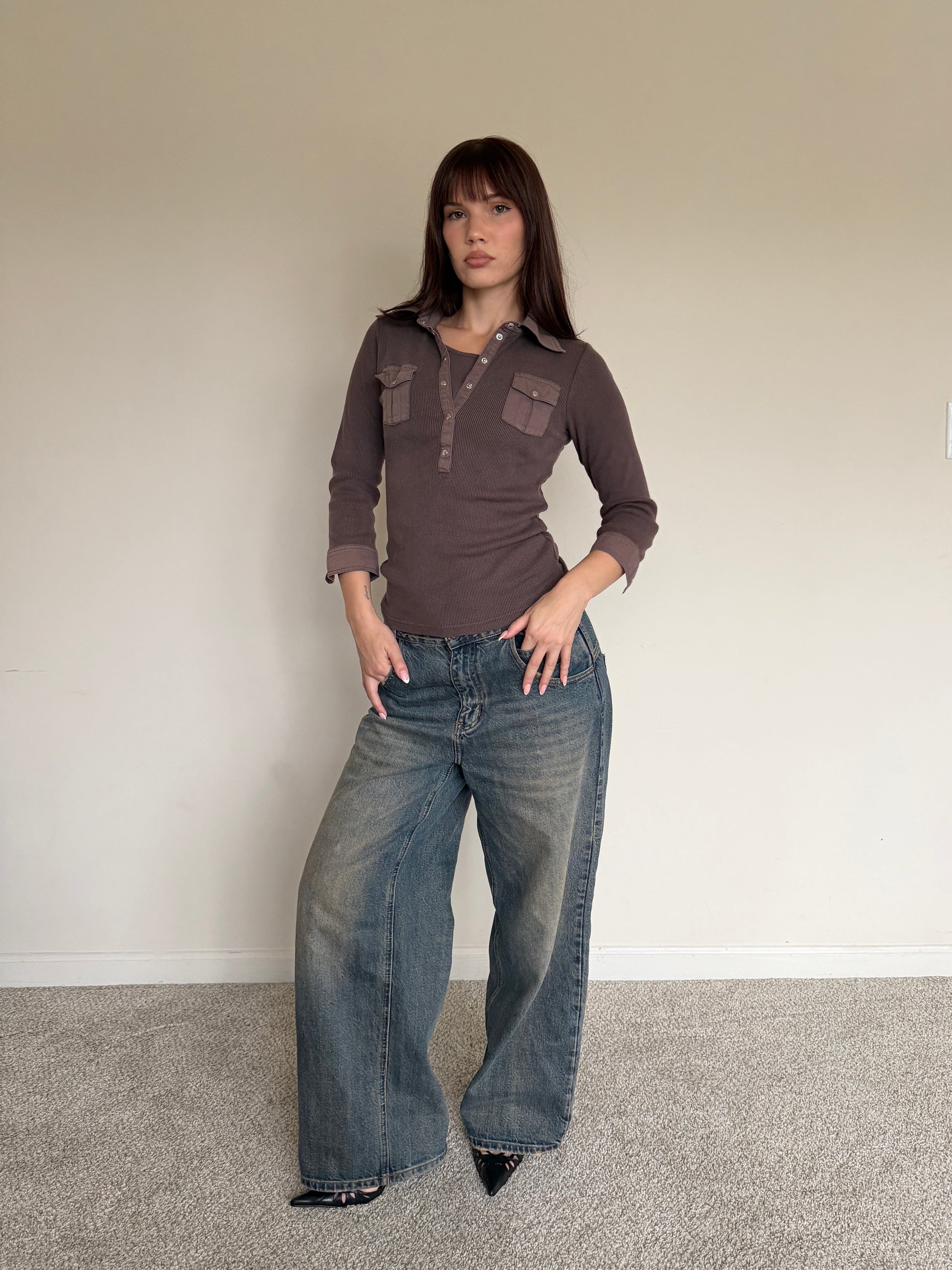 Woman wearing a brown shirt and blue jeans against a plain wall.