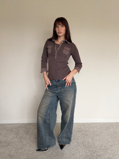 Woman wearing a brown shirt and blue jeans against a plain wall.
