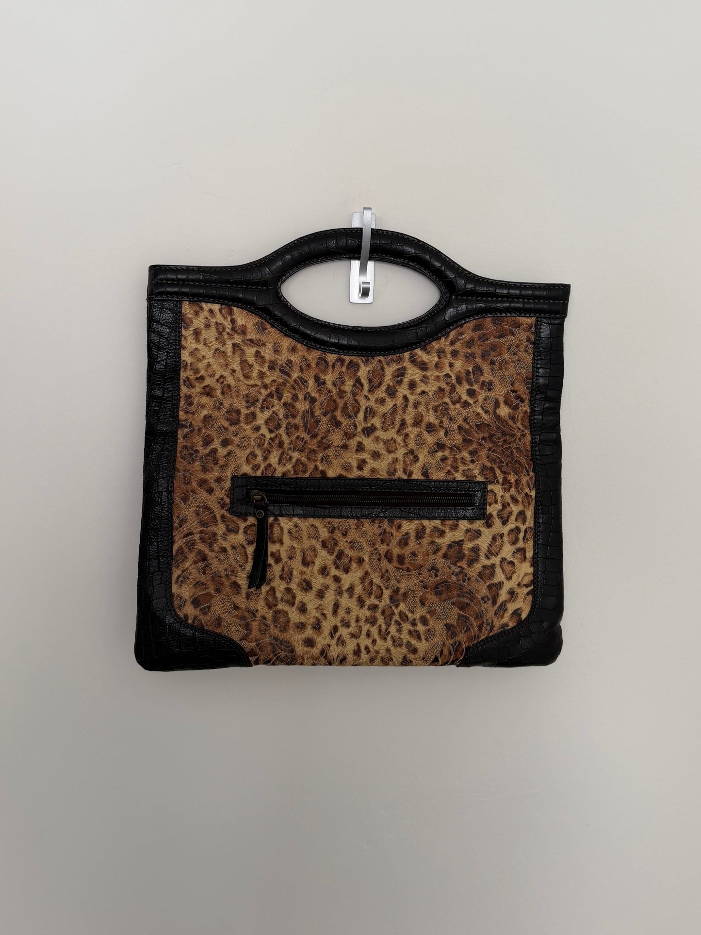 A leopard print handbag with black faux-leather trim and a top handle, hanging on a light gray wall.