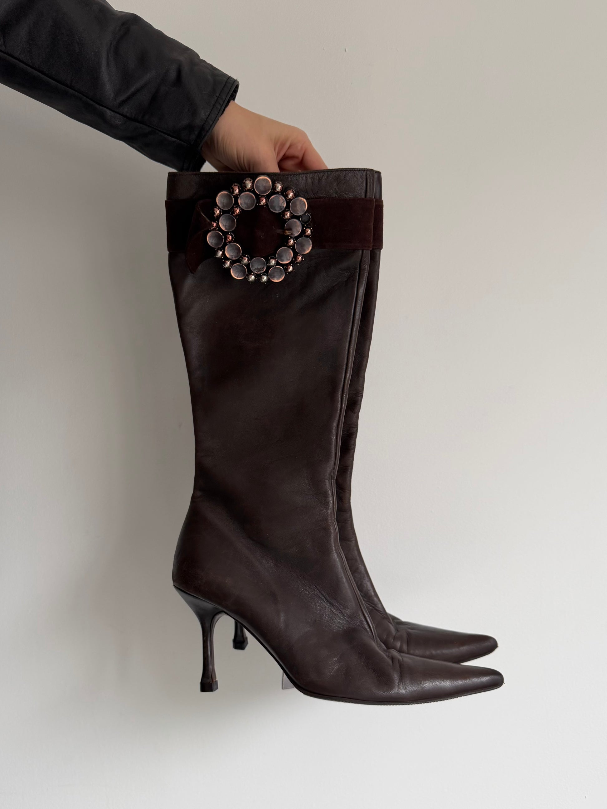 Black knee-high boot with decorative embellishment held by a person against a plain background