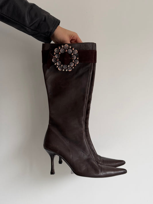 Black knee-high boot with decorative embellishment held by a person against a plain background