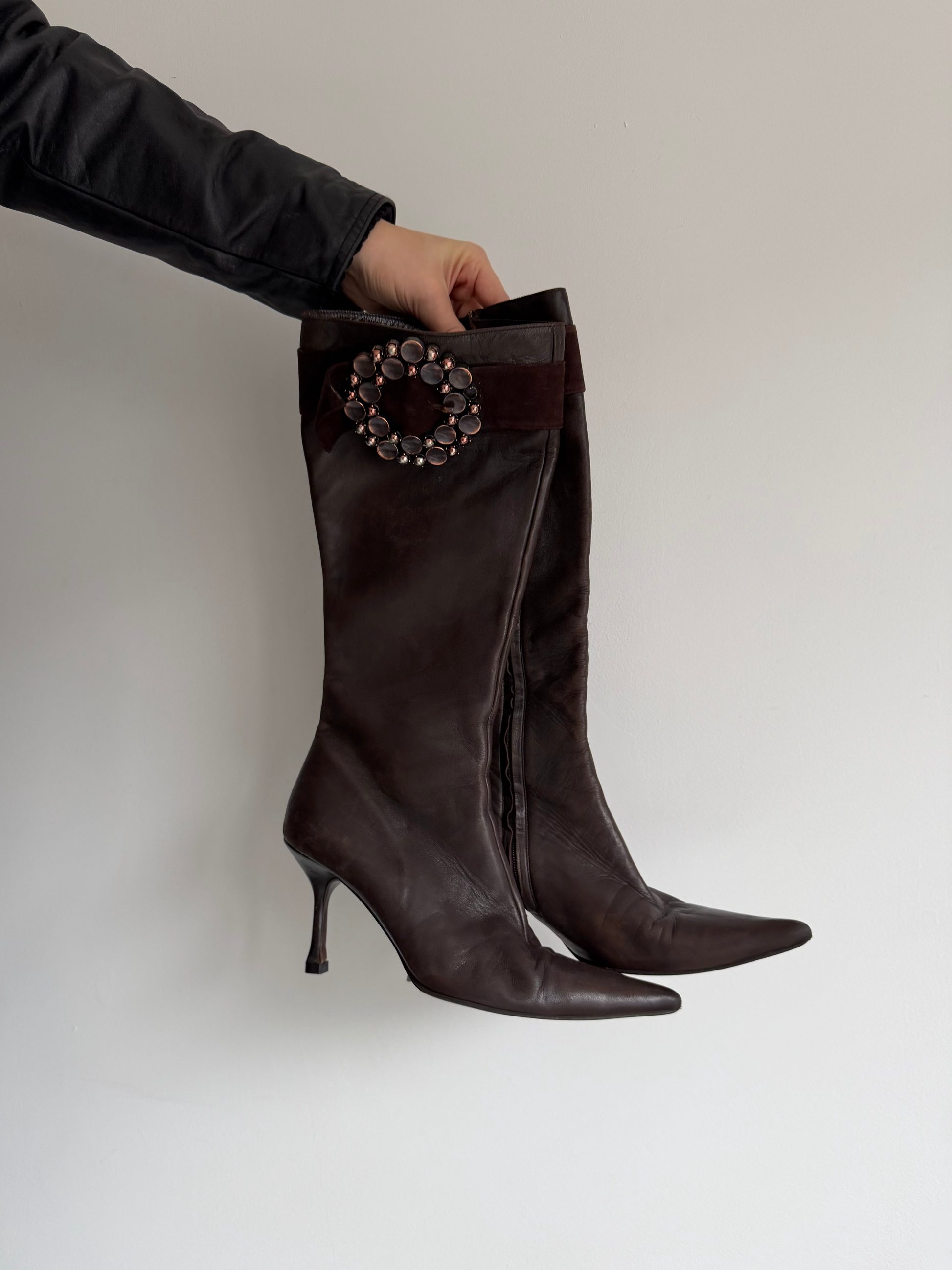 Brown knee-high boots with a decorative brooch on a light gray background
