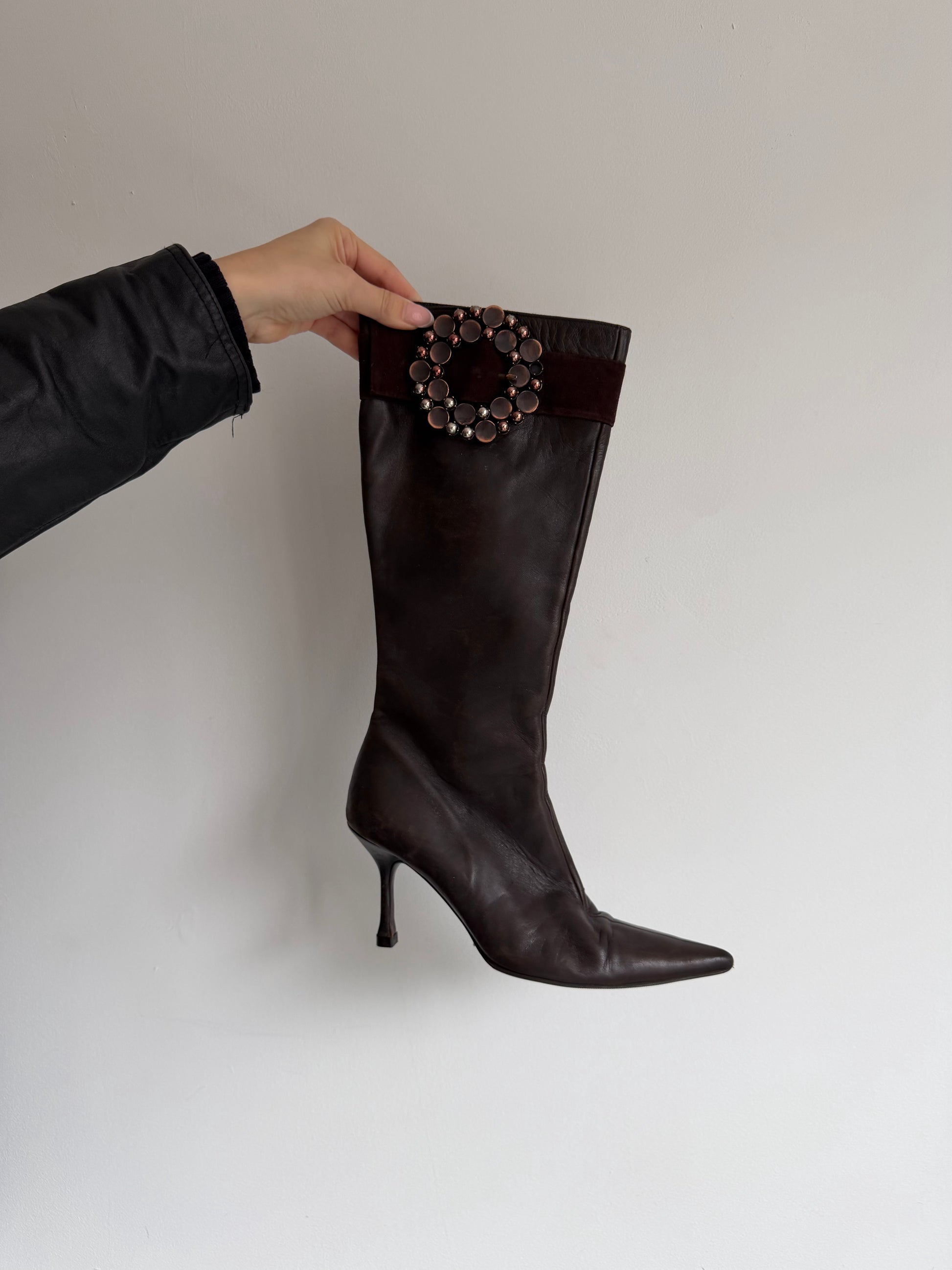 Brown knee-high boot with decorative embellishments held by a hand against a plain background