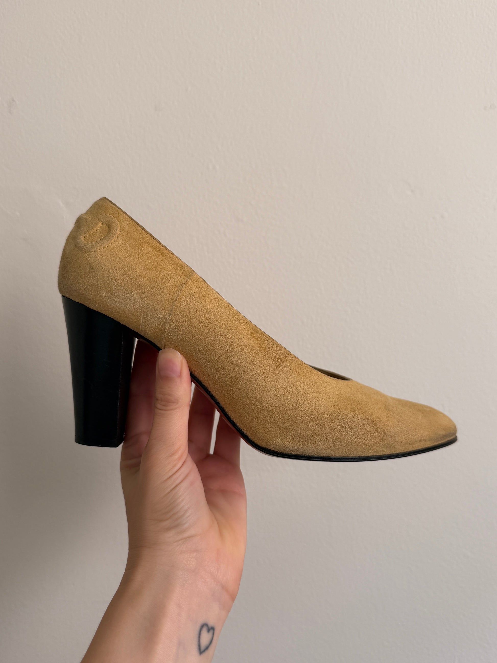 A hand holds a beige suede high-heeled shoe with a black block heel against a plain off-white wall.