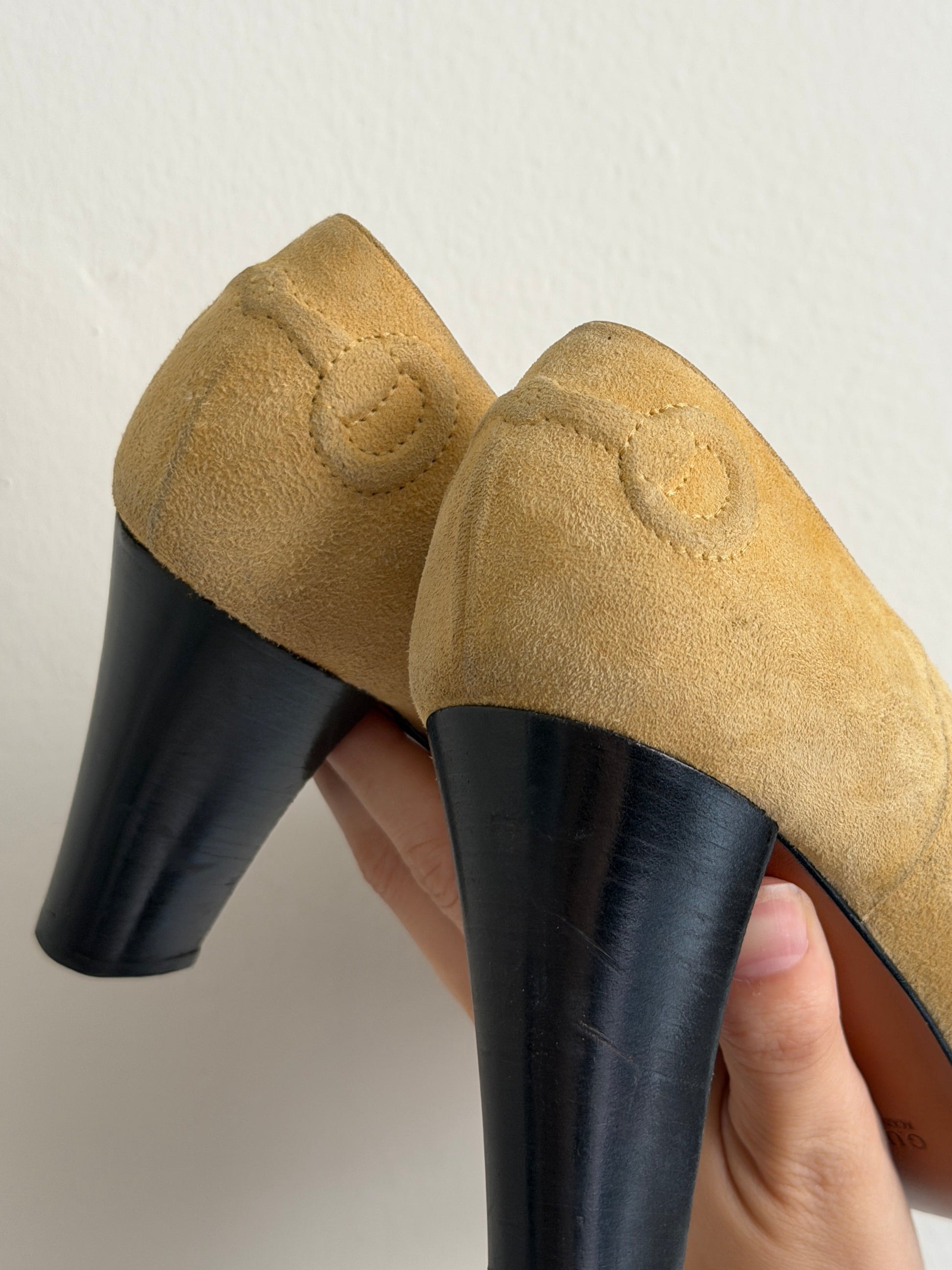 A pair of beige suede mule heels with black block heels, held by a hand against a plain off-white background.