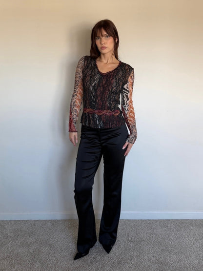 Person wearing a patterned top and black pants against a plain wall.