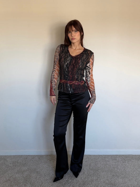 Person wearing a patterned top and black pants against a plain wall.
