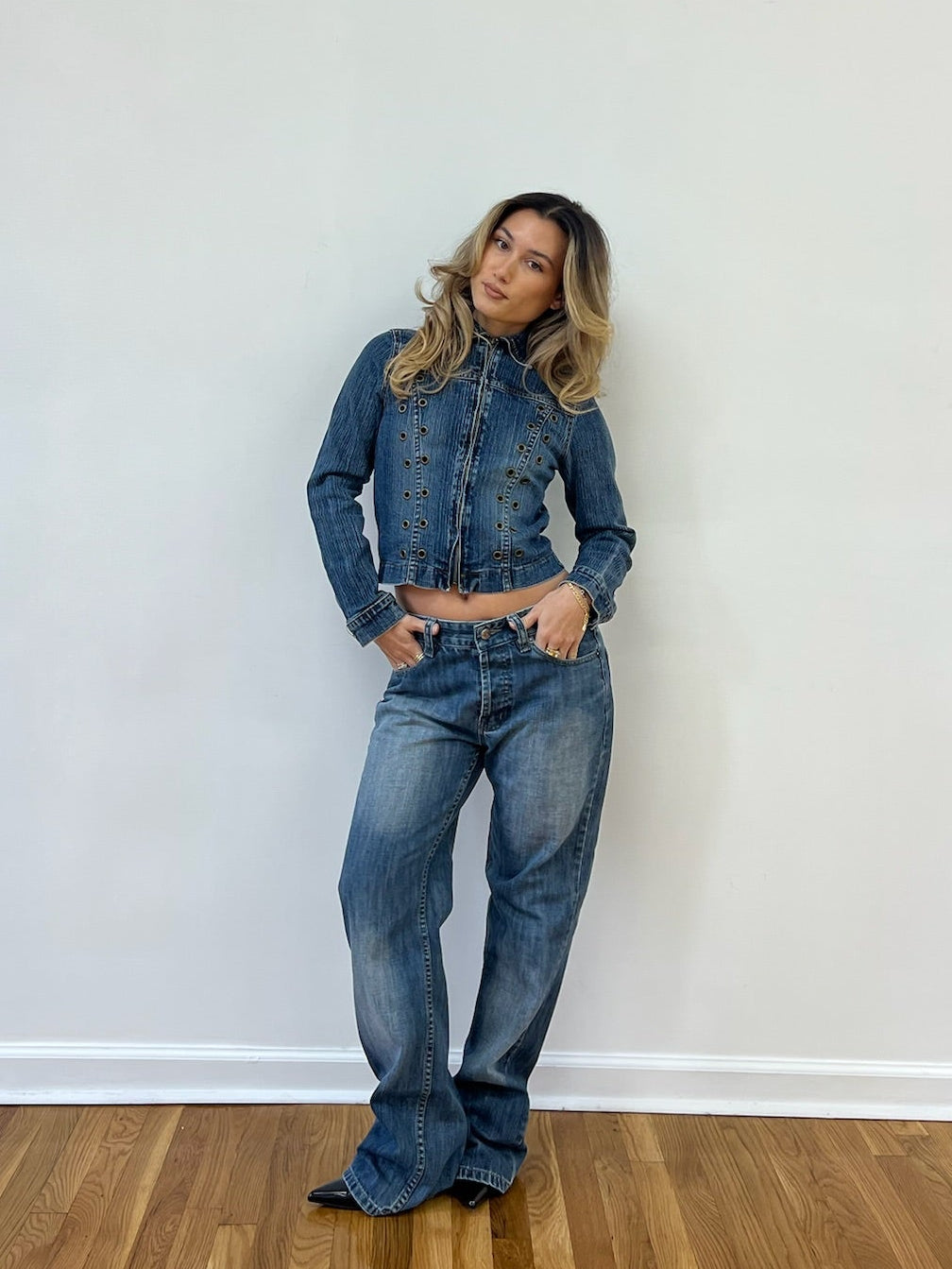 Woman wearing a denim jacket and jeans against a white wall.