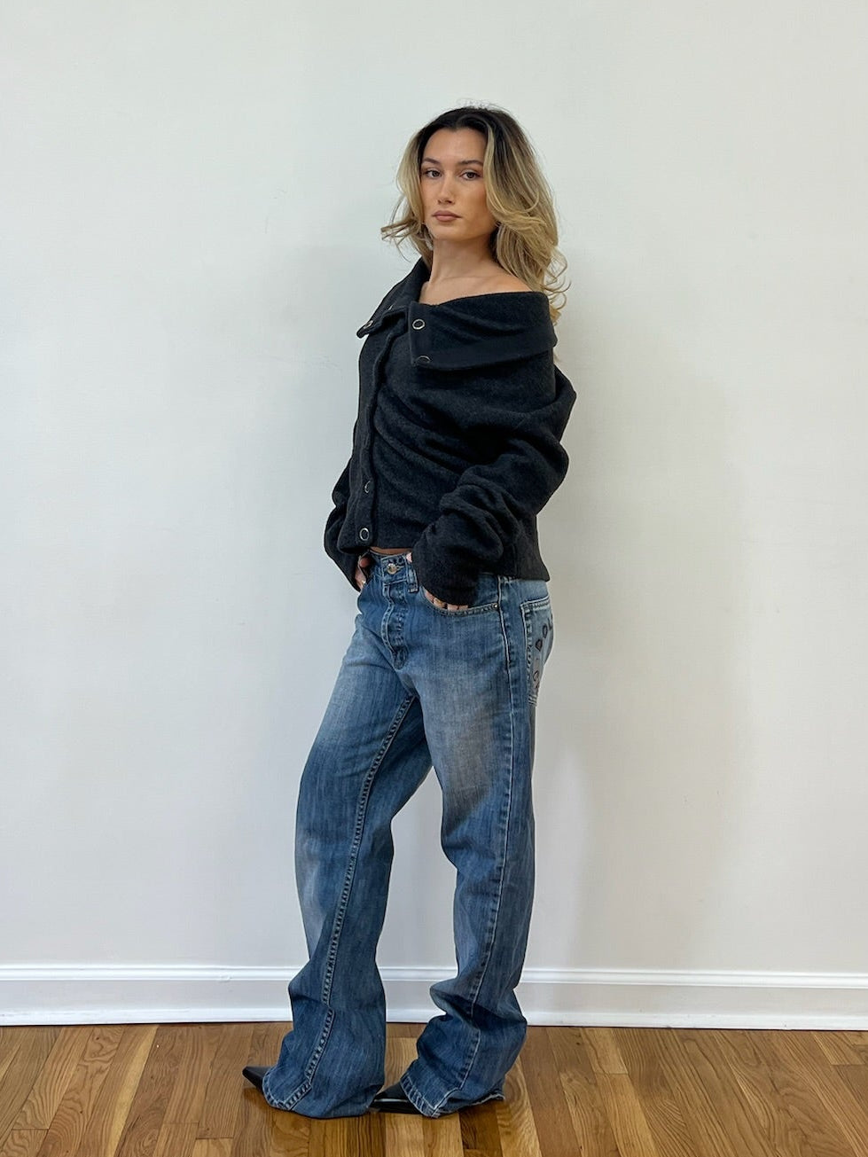 Person wearing a dark grey jacket and blue jeans against a white wall.