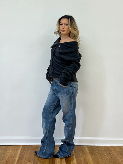 Person wearing a dark grey jacket and blue jeans against a white wall.